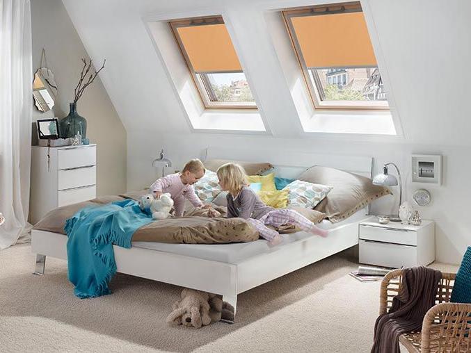 Two children play on the bed in the bedroom, the room is partly darkened by roller blinds on two wooden roof windows