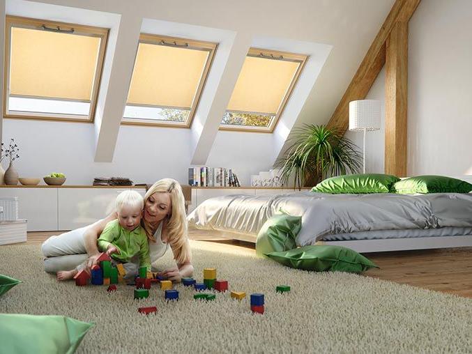 A mother plays with her child on the floor of the bedroom, in the background 3 wooden roof windows with almost closed roller blinds inside.
